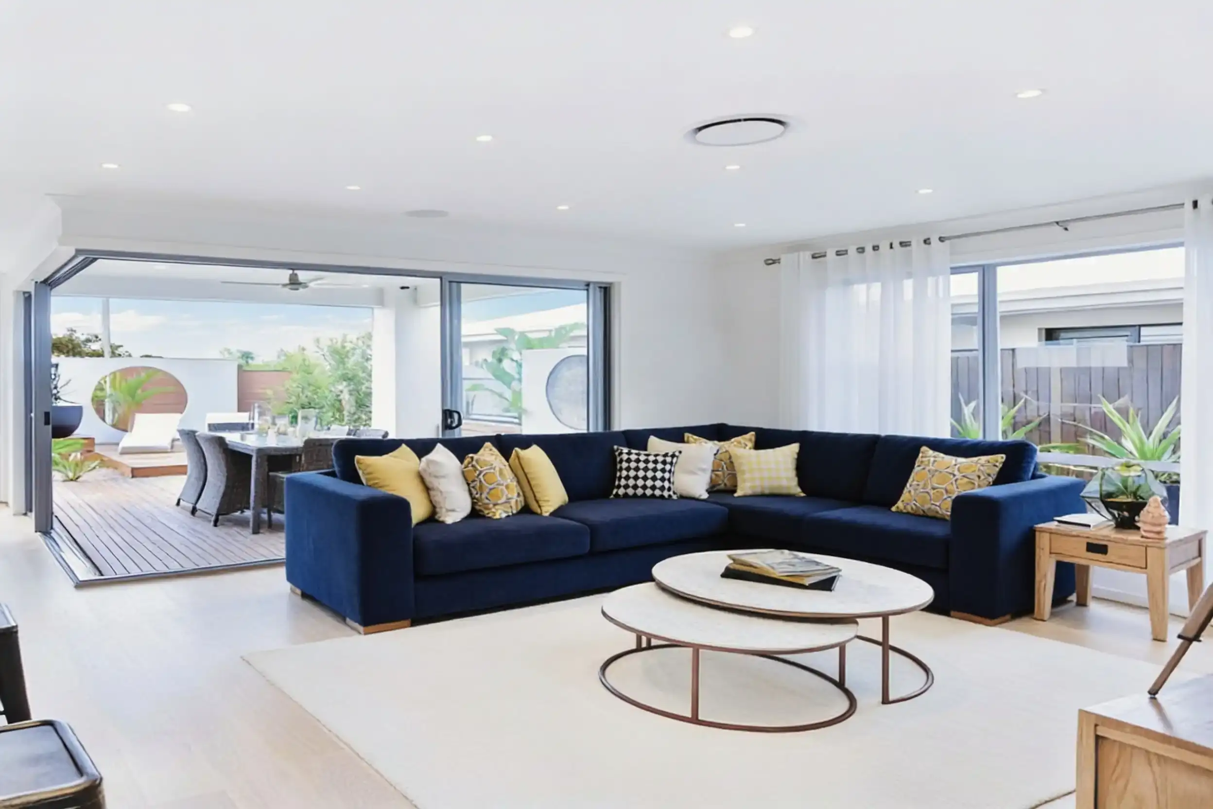 A bright open-plan living room with a round ceiling air vent above a navy corner sofa, glass doors open to a timber deck