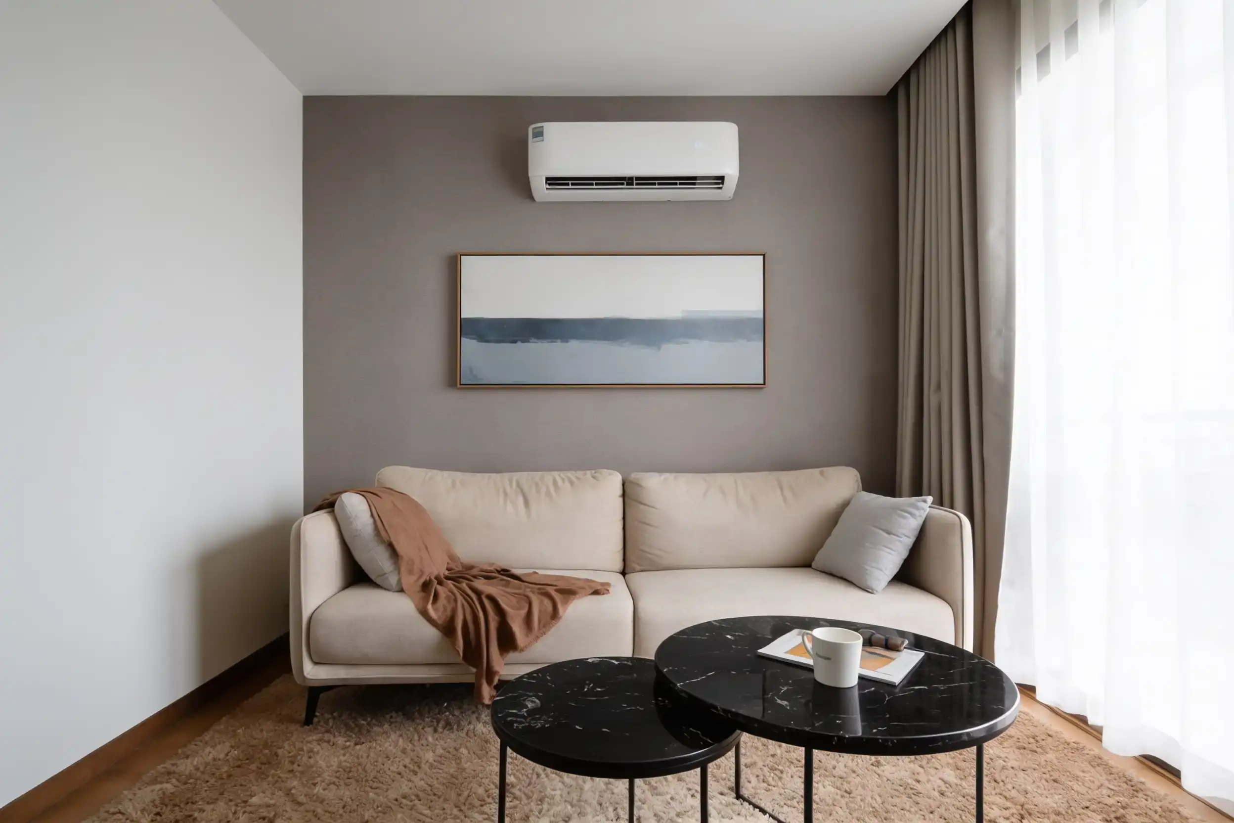 A white split system high on a taupe feature wall, above a framed print, a cream sofa with a rust throw and two black marble side tables
