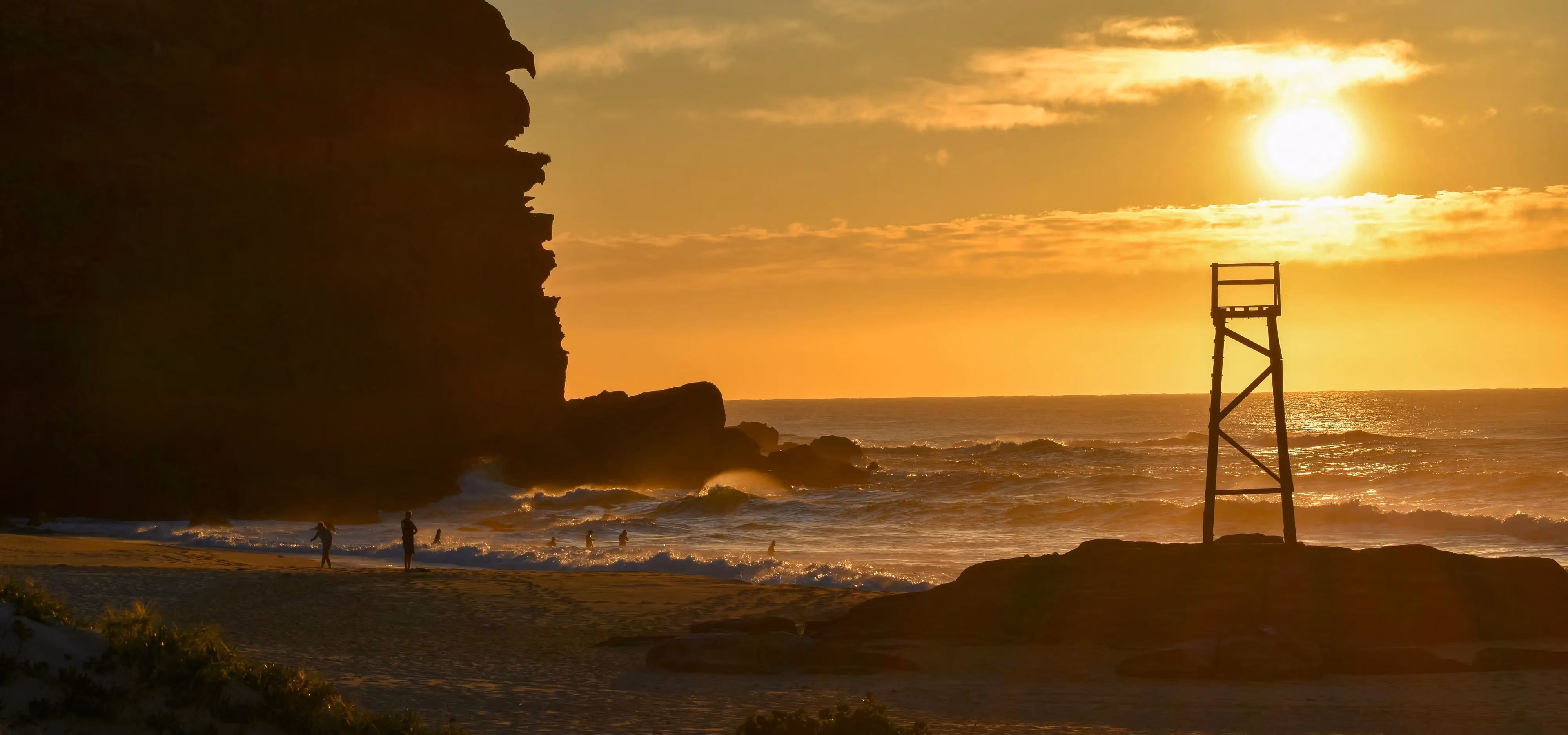 A timber shark lookout tower on a rock shelf and a dark headland, silhouetted against the low sun over the surf, swimmers in the waves