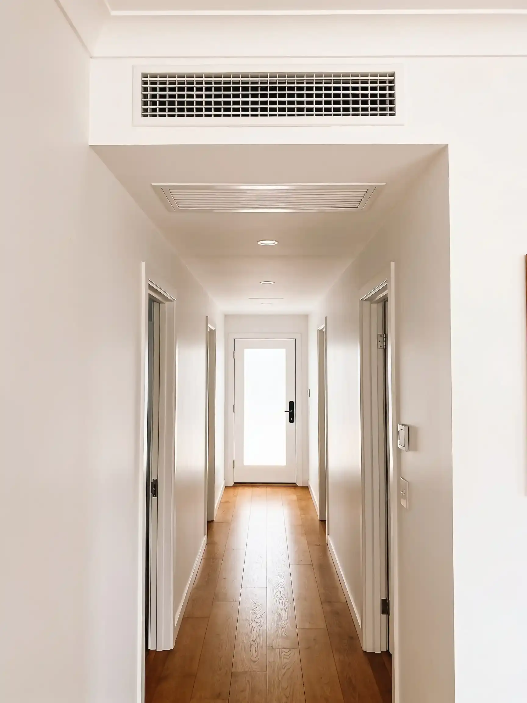 A long white hallway with timber floorboards, a return-air grille in the bulkhead and a linear outlet in the ceiling