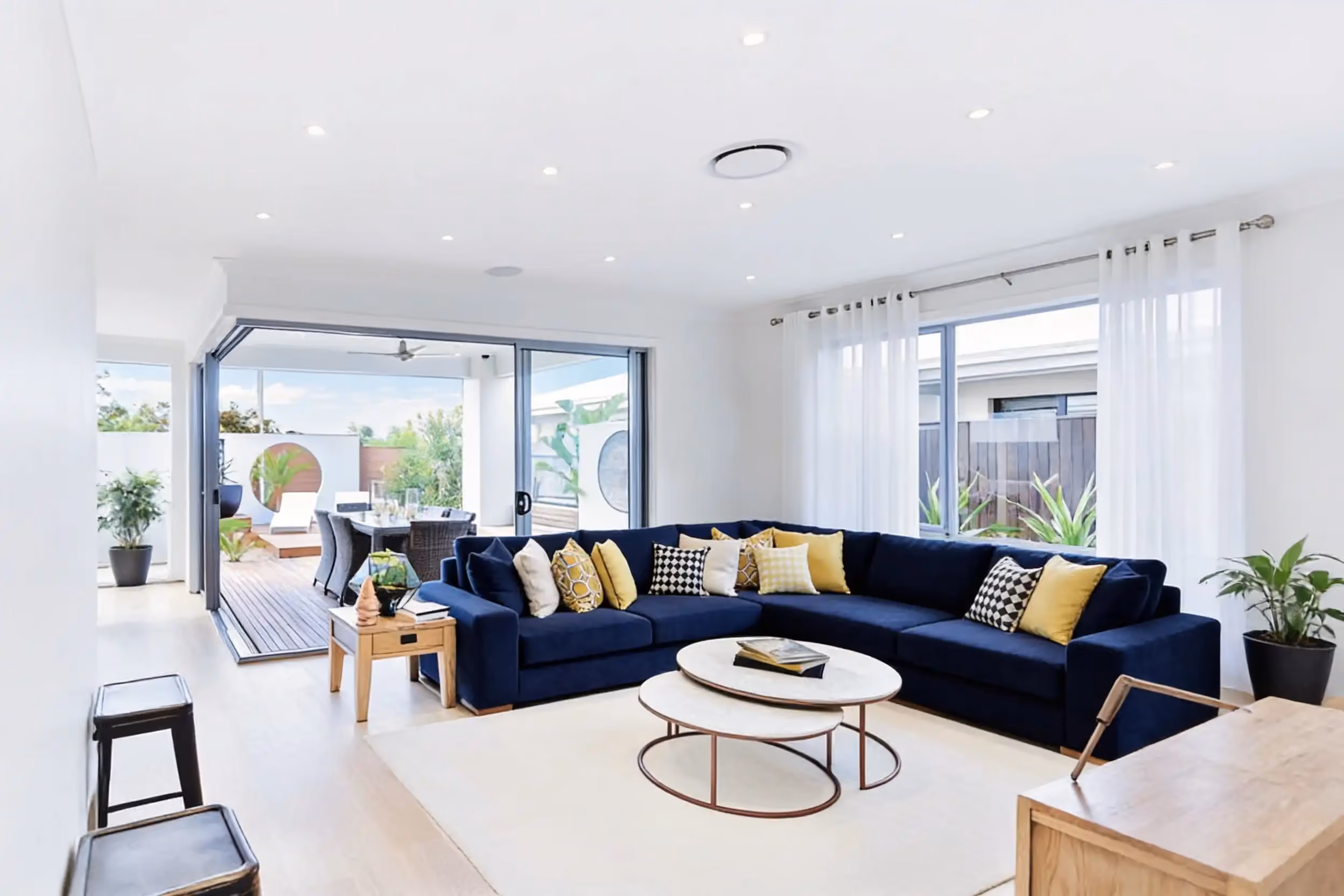 A bright open-plan living room with a round ducted air vent in the ceiling, a navy corner sofa and glass doors open to a deck